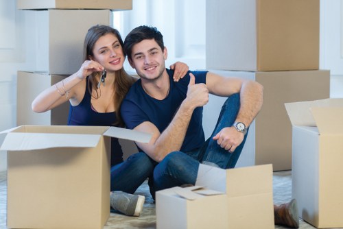 A young couple sitting on the floor inside a residential property surrounded by open cardboard moving boxes of various sizes, indicating a house move or relocation. The woman has long brown hair and is wearing a navy blue top, smiling and talking on a mobile phone while leaning against the man. The man has short dark hair, is wearing a dark t-shirt, and is giving a thumbs-up gesture with a cheerful expression. The background shows a bright, modern interior with light-colored walls and large windows allowing natural light to fill the room. The scene captures a moment of unpacking or preparing for a move, consistent with professional house removals services provided by Queens Park Man with Van in the London area, supporting local relocations in the postcode district of NW, near Queen’s Park, London.