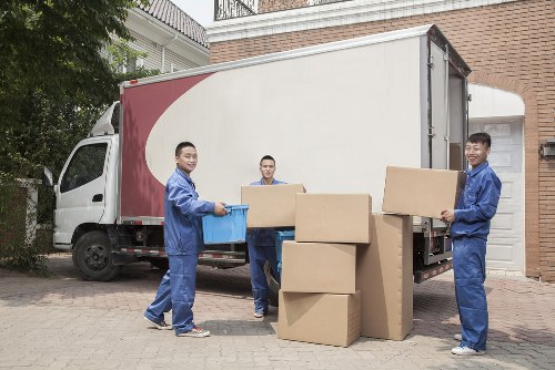 Three movers from Queens Park Man with Van, dressed in blue uniforms, are unloading cardboard boxes from a white moving truck parked on a residential driveway in a neighbourhood near London, with a brick house and a garden in the background. One of the movers is holding a small blue plastic container, while another is carrying a medium-sized box, and the third is holding a large rectangular box. Several other boxes of various sizes are stacked on the ground beside them, ready for transportation or placement inside the house. The scene is set during daylight, with natural light illuminating the area, and the background includes elements of a typical residential area in London, such as a brick wall and greenery, reflecting the local context of house removals services in the area.