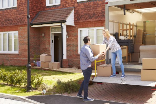 In front of a residential brick house in Queens Park, a woman with blonde hair wearing a striped long-sleeve top and blue jeans is seen loading a cardboard box into the back of a parked removals van. The van has its side door open, revealing interior shelving, wrapped furniture, and multiple packed cardboard boxes. A man with dark hair, dressed in a light grey jacket and dark trousers, stands on the driveway, reaching out to receive the box from the woman. Several additional cardboard boxes are stacked near the entrance of the house, which has a white front door and white-framed windows. The scene takes place during daylight with natural lighting, and surrounding the house is a small garden with green grass, shrubs, and a lamppost close by on the paved driveway, indicative of a suburban residential area nearby Queens Park postcodes.