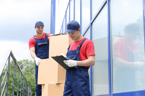 Two removal workers dressed in red T-shirts, navy overalls, and navy caps are seen outdoors beside a modern glass building, preparing cardboard boxes for a house move. The worker in the foreground is holding a clipboard and appears to be checking or recording items, while wearing white gloves. Behind him, the second worker is carrying two large cardboard boxes, one stacked on top of the other, with their natural brown surfaces visible and reinforced edges. The background reveals a partly cloudy sky and some trees, suggesting a residential or commercial property in the vicinity of Queens Park. The workers are positioned on a paved surface adjacent to a metal railing, with the glass wall of the building reflecting their figures. The scene captures a professional and organized atmosphere typical of house removal or relocation services provided by Queens Park Man with Van, emphasizing careful handling of packed household items in preparation for a local moving service within the London area.