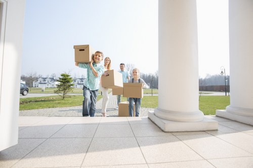 Four individuals, likely a family or removals team, are outside a white neoclassical building with large columns, preparing to load cardboard boxes onto a removals vehicle in a residential area. One person in a light blue checked shirt is smiling and holding a small cardboard box in the foreground, with two others behind holding larger boxes, and a fourth person further back also carrying a box. The boxes vary in size and are made of plain brown cardboard, with some appearing to contain household items. The scene features a paved area in front of the building, which has a grass lawn and small trees visible in the background. The sky is overcast but bright, suggesting daytime, and the setting appears quiet with a few parked cars and residential houses further away. This scene exemplifies house removal preparations, with the team efficiently sorting and carrying belongings, reflecting professional relocations in the Queens Park area.