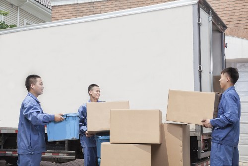 Three men dressed in blue work uniforms are unloading cardboard boxes and plastic storage containers from a white moving van parked on a residential street, possibly within the Queens Park or surrounding NW postcode area. The background features a brick house wall and a driveway, indicating a typical UK home environment. The men are carefully handling the items; one is carrying a blue plastic container, another is passing a cardboard box to a colleague, while the third man is holding a box ready for loading or unloading. The vehicle is positioned parallel to the curb, with its rear doors open, revealing the clean, spacious cargo area. The scene is well-lit, suggesting daytime outdoor conditions. Visible on the ground are additional cardboard boxes, with some stacked inside the van, demonstrating a systematic moving process. This image illustrates professional removals service personnel from Queens Park Man with Van engaged in careful, organized loading or unloading activities at a residential property, reflecting their expertise in house removals across NW London neighborhoods.