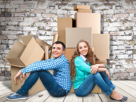 A young man and woman sit on light-colored wooden flooring in front of a stack of brown cardboard moving boxes of various sizes, some open and partially filled, against a weathered brick wall interior in a residential setting. The man, wearing a blue checkered shirt and dark jeans, has short dark hair and is smiling while leaning back slightly, supporting himself with one hand on the floor. The woman, dressed in a teal sweater and blue jeans, has long, curly reddish hair and is smiling brightly with her arms crossed over her knees. The boxes appear to be packed and ready for a house move, with some boxes slightly tilted or leaning against the wall. The environment suggests an active moving process, typical of a house removal in the Queen's Park or nearby London area, as indicated by the professional context of Queens Park Man with Van's removals services.