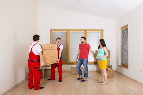 In a bright, empty room with white walls and light wooden flooring, two movers dressed in red overalls are carrying a wooden chest of drawers along with another mover, a man in casual clothing, and a woman in a blue top and yellow skirt. The movers are holding the furniture carefully as they prepare to load it into a vehicle or position it within the space. Behind them, three sizable windows with wooden frames allow natural light to illuminate the room, which appears to be part of a residential property in the Queens Park area. The room is free of additional furniture or clutter, emphasizing the process of house removals or furniture relocation. Several cardboard boxes are stacked in the corner on the right, indicating a move in progress. The environment is clean, well-lit, and empty, suitable for professional house removals and furniture transportation, exemplifying a typical scene in a household preparing for moving day. Queens Park Man with Van’s services are likely to facilitate such furniture handling and home clearance tasks efficiently.