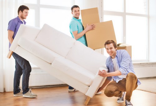 Three men are engaged in a house moving task inside a bright, spacious room with large white-framed windows allowing natural light to fill the space. The man in the foreground, wearing a light blue shirt and brown trousers, is crouching and holding the front corner of a large, cream-colored fabric-upholstered sofa, which is partially lifted off the ground, indicating effort in maneuvering the heavy furniture. Behind him, another man dressed in a purple t-shirt and dark jeans is helping to steady the sofa’s backrest as they work together. A third man, dressed in a turquoise polo shirt and dark trousers, is standing slightly further back, smiling while holding a closed cardboard box. Additional moving cartons and a small stack of cardboard boxes are visible on the wooden floor, suggestive of packing for a house move. The setting appears to be a residential interior, with a minimalistic and clean appearance, indicating a house or apartment in the Queens Park area, relevant to the services of Queens Park Man with Van, who specialise in removals and relocation within London and the surrounding postcode districts.