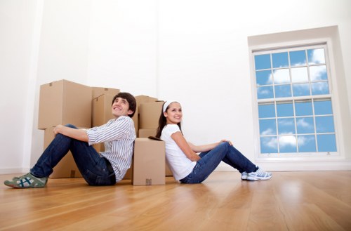 A young couple, a man and a woman, are seated on a polished wooden floor inside a bright, empty room with white walls. They are surrounded by several cardboard moving boxes in various sizes, some stacked against the wall and others placed on the floor around them. The man, wearing a light striped shirt and jeans, is leaning back with his legs extended and crossed at the ankles, smiling and looking upward. The woman, dressed in a white t-shirt and jeans, is sitting relaxed with her legs stretched out and hands resting on her knees, smiling at the camera. Behind them is a large, white-framed window with multiple panes, through which a clear blue sky and some fluffy white clouds are visible. The natural light from the window illuminates the room, emphasizing the wooden floor and the simplicity of the space, reflecting a typical residential setting suitable for house removals or moving services in the local area, such as Queens Park or nearby London neighborhoods. This scene appears to capture a moment of packing or unpacking, relevant to professional removals services like those offered by Queens Park Man with Van.