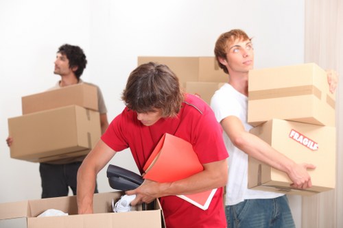 Three men involved in a house relocation process are visible inside a residential property, with a focus on moving boxes and personal belongings. The man in the foreground, wearing a red T-shirt, is leaning over a large cardboard box, holding a pair of white sneakers and a black handheld device, possibly a mobile phone, with a red folder or binder tucked under his arm. Behind him, another man with light brown hair, dressed in a white T-shirt, is carrying a medium-sized cardboard box labeled 'Fragile,' with a focused expression on his face. To the left, a third man with curly dark hair, wearing a grey T-shirt, is carrying a large cardboard box against his chest, looking towards the left side of the image. The background suggests an indoor setting, with a plain white wall and possibly a doorway or corridor. The scene is well-lit, highlighting the textures of the cardboard boxes, the different fabric finishes of the clothing, and the household items being moved, all indicative of a professional removals service operating in the Queens Park area. Queens Park Man with Van is involved in assisting with house moves, as reflected in the careful handling of household items and organized scene.