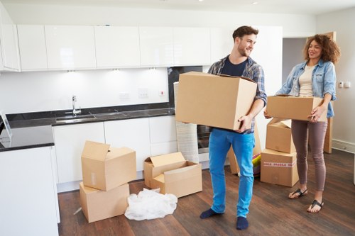 A man and a woman are inside a modern, well-lit kitchen with white cabinetry and a black countertop, preparing to move boxes. The man, dressed in a checked shirt and blue jeans, is holding a large cardboard box close to his chest, smiling as he stands on a dark wooden floor. Several smaller cardboard boxes, some open and others sealed, are scattered on the floor in front of him, along with crumpled packing paper. The woman, wearing a light denim jacket over a patterned top and beige trousers, stands nearby holding another medium-sized box, smiling towards the man. In the background, the kitchen wall features white cabinets and a window allowing natural light to illuminate the scene, creating a professional yet casual atmosphere typical of a house removal in the Queens Park area. The setting emphasizes a domestic environment suitable for local removal services offered by Queens Park Man with Van, illustrating the process of packing and moving within residential properties in the London NW10 postcode district.