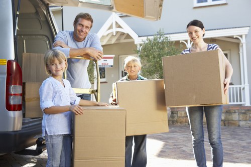 A family of four, including two young girls and a woman, are standing outside a residential house in a suburban area of Queens Park, with a removals van parked on the driveway. They are holding several large cardboard boxes, which are used for house packing and moving. The woman is on the right side of the image, wearing a striped top and jeans, smiling while holding a box. The man, on the left, is leaning slightly on the open van door, dressed in a light blue shirt, also smiling. The two children, a girl on the far left and a boy next to her, are each holding boxes, both dressed casually in light tops and jeans, smiling at the camera. Behind them, a glimpse of the house with a front porch and some greenery is visible, indicating a typical UK residential setting. The scene takes place during daylight with clear weather, suggesting a planned house removal by Queens Park Man with Van, a professional removals service covering the local area of Queens Park and NW10 postcode district. The image captures the organized process of packing and loading household belongings for a residential move.