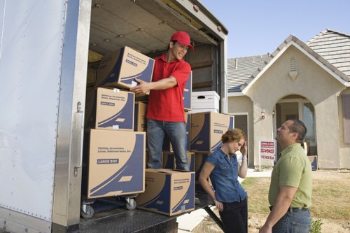 A professional removal worker, dressed in a red shirt and red cap, is inside the back of a large moving van, lifting and arranging cardboard boxes labeled for storage or relocation. The boxes are made of brown corrugated cardboard with blue and white printed labels, and some are stacked on a dolly or hand truck. Outside a residential property in a suburban area, a man and woman are standing on the driveway, discussing the move; the man is wearing a green polo shirt and glasses, while the woman has a blue button-up shirt with her hair tied back. In the background, a modern, pitched-roof house with a white exterior, grey roof tiles, and a small front garden with grass and a pathway are visible. Natural daylight illuminates the scene, highlighting the outdoor environment typical of house removals in the UK, subtly linking the service to a local area such as Queens Park or nearby suburbs. The setup indicates an ongoing or completed part of a residential move, with focus on household items being carefully packed and loaded for transportation by Queens Park Man with Van, a specialist in removals services.