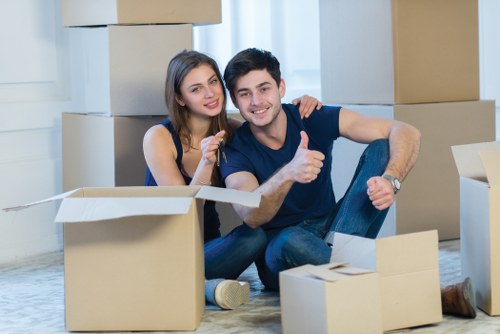 A young couple with light skin and dark hair sit on the floor inside a residential property, surrounded by open cardboard boxes of various sizes. The woman is wearing a black sleeveless top and smiling while pointing at the camera; the man, dressed in a navy T-shirt and jeans, is giving a thumbs-up gesture. Behind them, large stacks of sealed cardboard boxes rest against a white wall, indicating they are in the process of packing or unpacking. The environment suggests a typical house interior with natural light streaming through a window, illuminating the scene. The couple appears to be preparing for a house move, consistent with a professional removals service such as Queens Park Man with Van, servicing areas around the NW postcode area in London, including Queens Park. The setting is clean, organized, and indicative of a household transition, with no visible furniture or appliances in view, just packing materials and personal belongings in transit during a house relocation process.