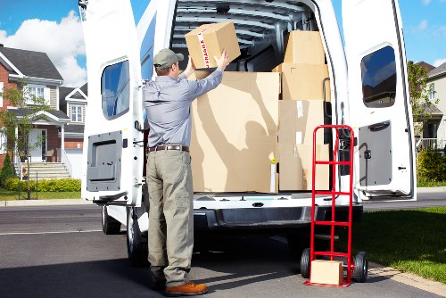 A man wearing a grey shirt, beige trousers, and a grey cap is standing at the rear of a white removals van with its back doors open, loading or unloading several cardboard boxes of various sizes. The boxes are brown with minimal labeling and are positioned both inside the van and in his hands. A red hand truck with black wheels is leaned against the side of the van, holding one cardboard box on the bottom. The van is parked on a residential street in a suburban neighbourhood with well-maintained modern houses, some with brick facades, visible in the background. The scene is set during daylight hours with a partly cloudy sky, and the environment suggests a house relocation, possibly within or near queenspark, where Queens Park Man with Van provides removals services. The focus on household items like cardboard boxes and the loading activity aligns with professional house removals and relocation activities common in London suburbs.