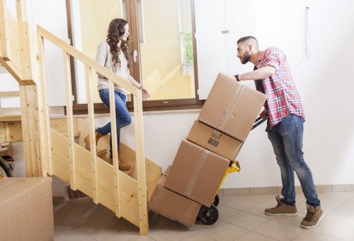 A man wearing a red and white checked shirt, jeans, and grey shoes is using a hand truck to transport three cardboard boxes stacked on top of each other, towards a staircase in a residential home. He is guiding the boxes up the small wooden staircase with a safety strap attached to the trolley for stability. A woman with long, curly brown hair, dressed in a white top and blue jeans, is sitting at the bottom of the staircase, watching the process. The interior space has light-colored walls, a side window letting in natural light, and tiled flooring. Nearby, there is additional packing material, including more cardboard boxes, indicating a moving or packing scenario, likely part of a house removal service in Queens Park, London, provided by Queens Park Man with Van. The scene captures a typical moment during a local house removal, focusing on careful manual handling of household items in a domestic environment.