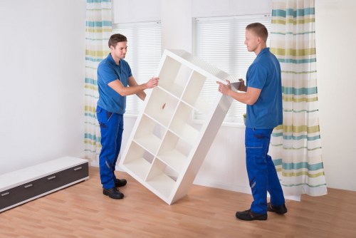 Two movers from Queens Park Man with Van, dressed in matching blue uniforms, are carefully lifting a white wooden bookshelf with multiple compartments in a residential interior. The bookshelf has a rectangular shape with a simple, modern finish, and is being positioned upright between the two men. The room has a light wooden floor, and in the background, there are three large windows with white blinds and striped curtains in shades of blue, green, and cream, allowing natural daylight into the space. A long white radiator is affixed along the wall near the floor on the left side of the image, and the overall setting suggests an ongoing house removal or interior reorganization. The scene highlights a professional, careful handling of household furniture typical of a house removals service, accurately depicting the process of moving items in a domestic setting, with subtle contextual clues pointing to a UK home environment, possibly within or near London, such as Queens Park, NW, from the postcode area inferred from the webpage title. The focus is on safe, methodical transport of furniture as part of professional removals.