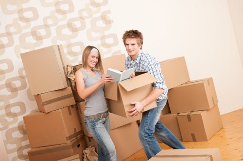 A young man and woman are inside a residential room with a light-colored, patterned wallpaper and wooden flooring, surrounded by multiple large cardboard moving boxes of various sizes, some stacked and others placed on the floor. The woman, wearing a grey sleeveless top and jeans, is holding an open book or magazine and smiling, while the man, dressed in a plaid shirt and jeans, is in the process of lifting or arranging a box with a focused expression. The background includes a plain, neutral-colored wall with a repeating geometric pattern and a slightly visible door or opening to another room. The scene appears to depict a house relocation, possibly in a UK town, with the pair actively involved in packing or unpacking, supported by Queens Park Man with Van's moving services, reflecting a typical household moving scenario with neatly packed boxes and personal belongings ready for transportation.