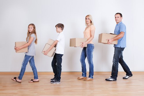 A family of four, consisting of a young girl, a boy, a woman, and a man, are engaged in a house removal process inside a bright, modern residential space in Queens Park with a light wooden floor and plain white wall background. The girl and boy, both dressed casually in jeans and T-shirts, are carrying cardboard boxes; the girl holds a medium-sized box, while the boy carries a slightly larger one. The woman, also in casual attire with blue jeans and a peach top, holds a medium-sized box, and the man, dressed in a blue shirt and dark jeans, carries a larger cardboard box. They are all walking in a line, actively engaged in loading or unloading, part of a professional removals service by Queens Park Man with Van. The shelves and furniture are minimal or not visible, focusing on the family members and their task, illustrating a typical move within a residential property in the area, emphasizing careful handling of household belongings during the relocation process.
