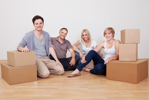 A group of four people, two men and two women, sitting on a light wooden floor in a bright, plain room, surrounded by several cardboard moving boxes of various sizes. The individuals are smiling and appear relaxed, with some leaning on or sitting beside the boxes. The boxes are sealed and unmarked, suggesting they are packed for a house move. The background features a white wall with no other visible furniture or decor, indicating an indoor residential setting in the UK, possibly near Queens Park, London. The image depicts a typical scene of a household preparing for relocation, highlighting the services of Queens Park Man with Van, a local removals company. The natural lighting emphasizes the casual, friendly atmosphere of the scene, suitable for a webpage about pricing and quotes for house removals in the local area.