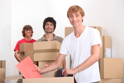 A group of three young adults in a bright, indoor residential space during a house move, surrounded by moving boxes of various sizes made of brown cardboard, some sealed with packing tape. The young man in the foreground, with light brown hair and wearing a white t-shirt, is smiling at the camera while placing a pink envelope into an open cardboard box. Behind him, a woman with dark blonde hair, dressed in red, and a man with dark curly hair, both wearing casual clothing, are holding additional boxes and smiling. The background shows a plain, light-colored wall with other stacked boxes, indicating an active moving process typical of homeowners or professional removals in a London suburb or nearby town. The environment is well-lit with soft natural or artificial light, emphasizing the organized clutter of household belongings preparing for transportation by a local removals service such as Queens Park Man with Van.