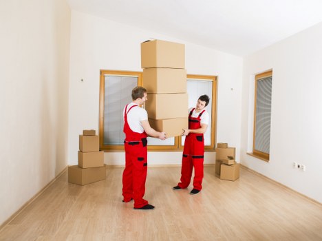 Two removal workers wearing red uniforms with black accents are inside a bright, empty room with white walls and light wooden flooring, engaged in stacking or rearranging large, flattened cardboard boxes. One worker is holding a tall stack of boxes while the other is assisting, both focused on the task. The room features two visible windows with wooden frames, one on each side, letting in natural light. Several smaller cardboard boxes are placed on the floor around them, some stacked near the windows. The setting appears to be a residential property in a suburban area, as suggested by the indoor environment consistent with house removals in the town. Queens Park Man with Van is known for providing professional removals services, and this scene depicts their team preparing to move or reorganise household items in a clean, unfurnished room. The clear, well-lit space emphasizes efficiency and care in the handling of household belongings during a move.
