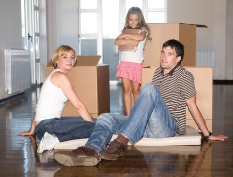 A family of three with light skin are positioned in a spacious, well-lit room with wooden flooring, likely during a house move. The mother, with shoulder-length blonde hair, is seated on the floor to the left, wearing a white sleeveless top and blue jeans, with her legs extended and hands resting on the floor behind her. The father, with short dark hair, is also seated on the floor, propped up on his hands, wearing a striped shirt and blue jeans, with brown shoes. They are surrounded by several open-top cardboard boxes, some placed upright, indicating packing or unpacking activities typical of a relocation service. Behind them, a teenage girl with long blonde hair, dressed in a pink skirt and white top, stands with her arms crossed, positioned near a stack of larger cardboard boxes, suggesting a family moving process. The background features large windows with frosted glass panes, radiating natural light into the room, and white radiators beneath the windows. This setting reflects a typical interior scene during a house removal, with a focus on household furniture, packed belongings, and the familial atmosphere associated with local house removals services in the UK, particularly in the vicinity of Queens Park or London NW, supporting the website’s SEO on recycling and sustainability within moving logistics provided by Queens Park Man with Van.