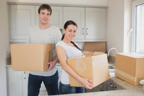 A young man and woman with light skin are in a modern kitchen preparing for a move, with the woman holding a medium-sized cardboard box filled with household items. The man stands slightly behind her, holding another box, both smiling and looking at the camera. The kitchen features light grey cabinets, a white countertop, and a stainless steel faucet. Several packed cardboard boxes are arranged on the counter and nearby, some sealed with packing tape, ready for transportation. Natural light streams through a window on the right, illuminating the scene and highlighting the clean, contemporary environment. The scene captures the process of household relocation, with furniture and belongings packed efficiently, underlining professional house removals services. Subtle signs of a residential property in the [POSTCODE] area suggest a home in [TOWN], where Queens Park Man with Van provides reliable and careful removals. The image emphasizes preparation, organization, and the human aspect of a smooth moving experience in a UK suburban setting, typical of local house relocation activities.