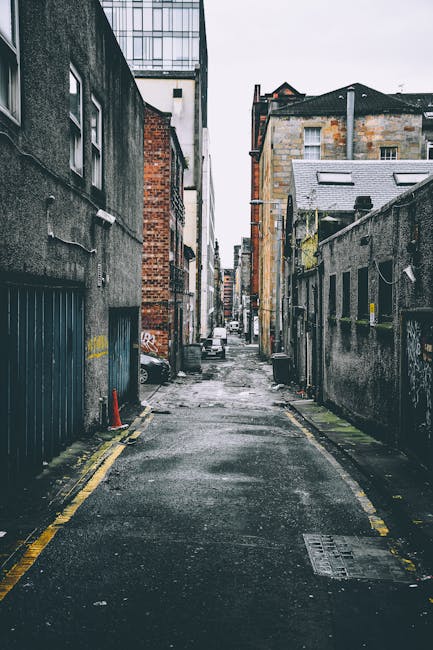 A narrow urban back alleyway with a wet, asphalt surface, flanked by tall, weathered brick and concrete buildings on both sides. On the left, there is a dark blue metal garage door with a safety cone 