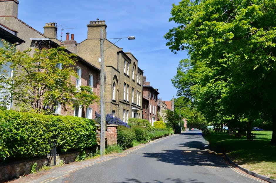 A narrow urban back alleyway with a wet, asphalt surface, flanked by tall, weathered brick and concrete buildings on both sides. On the left, there is a dark blue metal garage door with a safety cone 